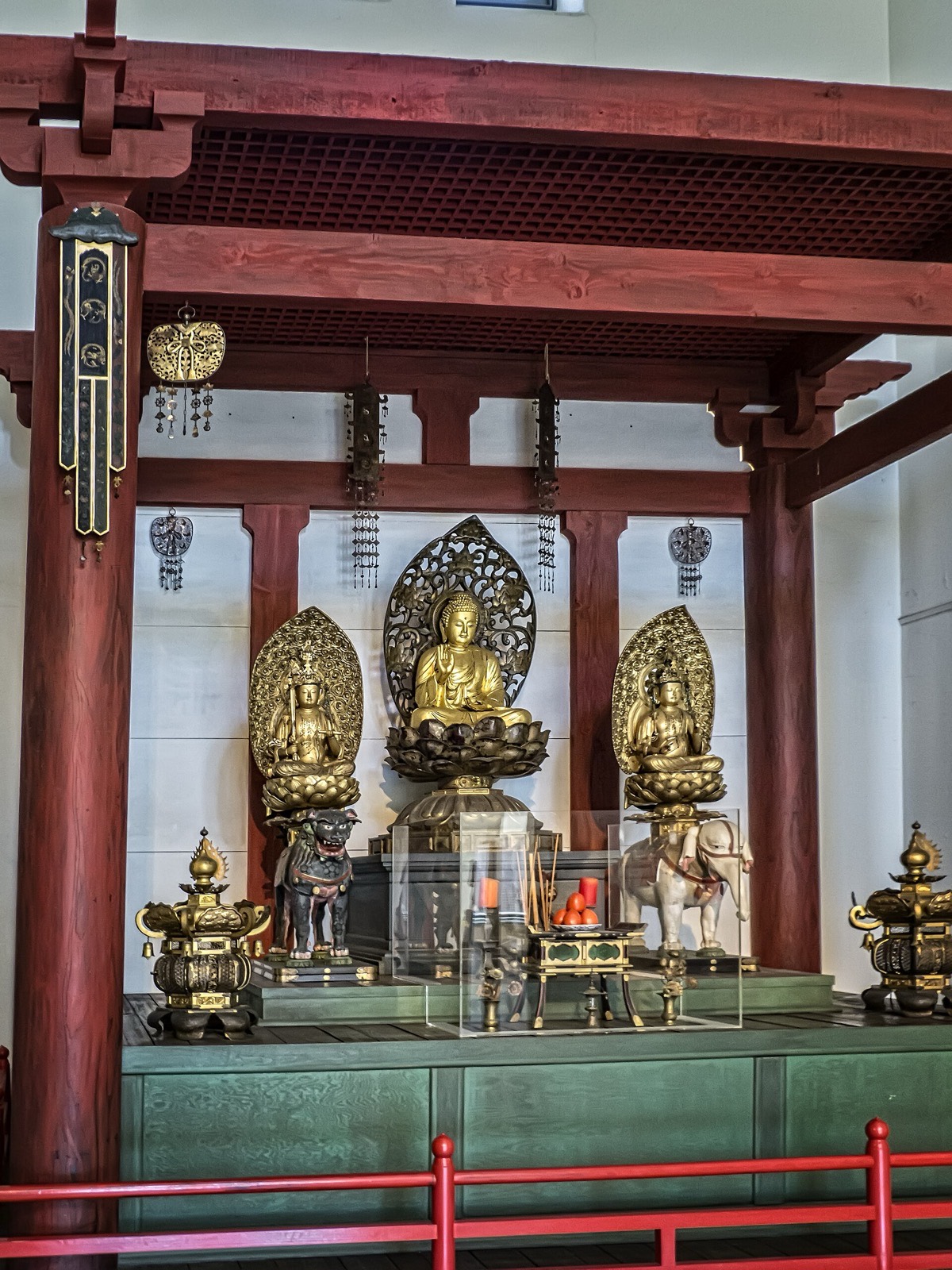Ruang dalam sebuah kuil Buddha Shingon: tiga arca Buddha berlapis emas di atas takhta teratai di bawah tiang-tiang merah, dengan sepasang lilin pilar oranye, dupa dan buah persembahan di meja di depannya.